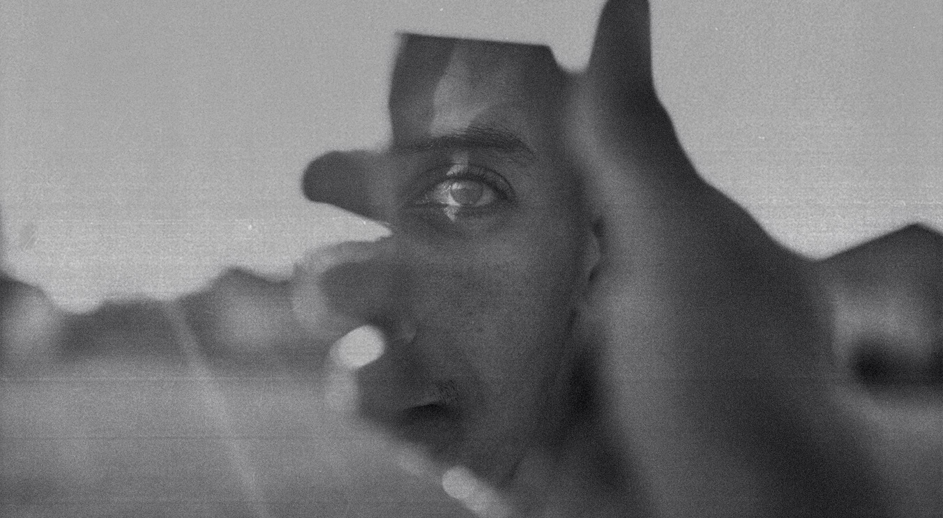 A black-and-white textured photo of a person holding up a shard of mirror, which is in focus and shows the reflection of their eye.