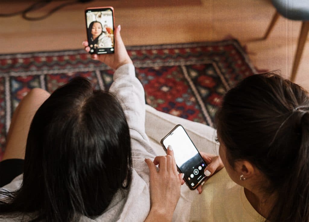 Two Asian girls sit on a couch, taking selfies.