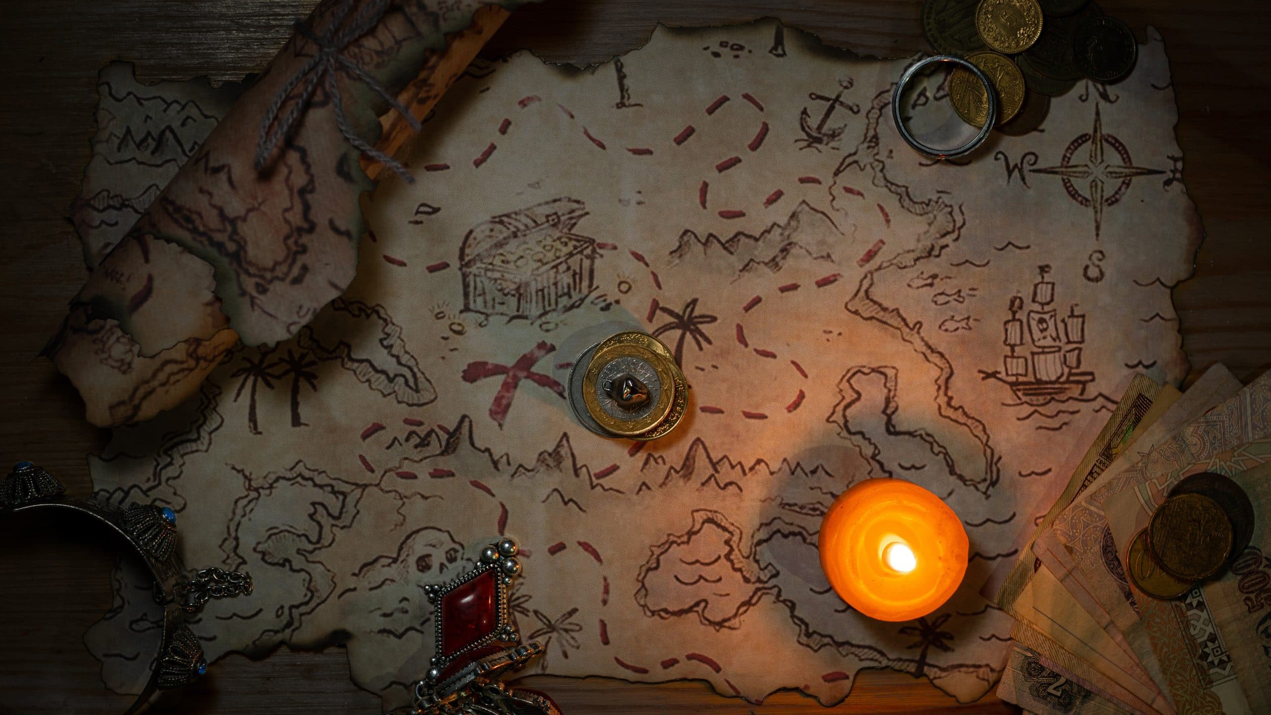 A treasure map laid out with a candle and some random articles splayed on top of it including coins, bills and some jewellery. It's a birds-eye view look at everything and it's dimly lit, and the map takes up a majority of the space in the image.