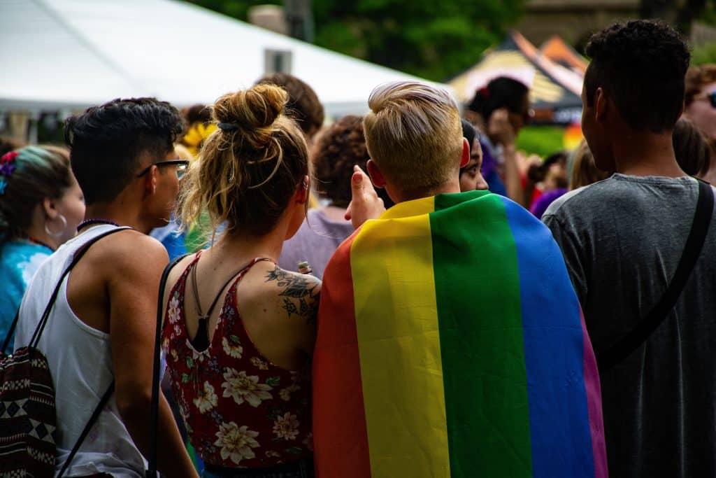 Several people are standing in very crowded area facing away from the camera. The age range seems to be quite young - people in their late teens or twenties, and they're wearing various clothing, however, one person stands out wearing a rainbow flag draped behind their back like a cape.