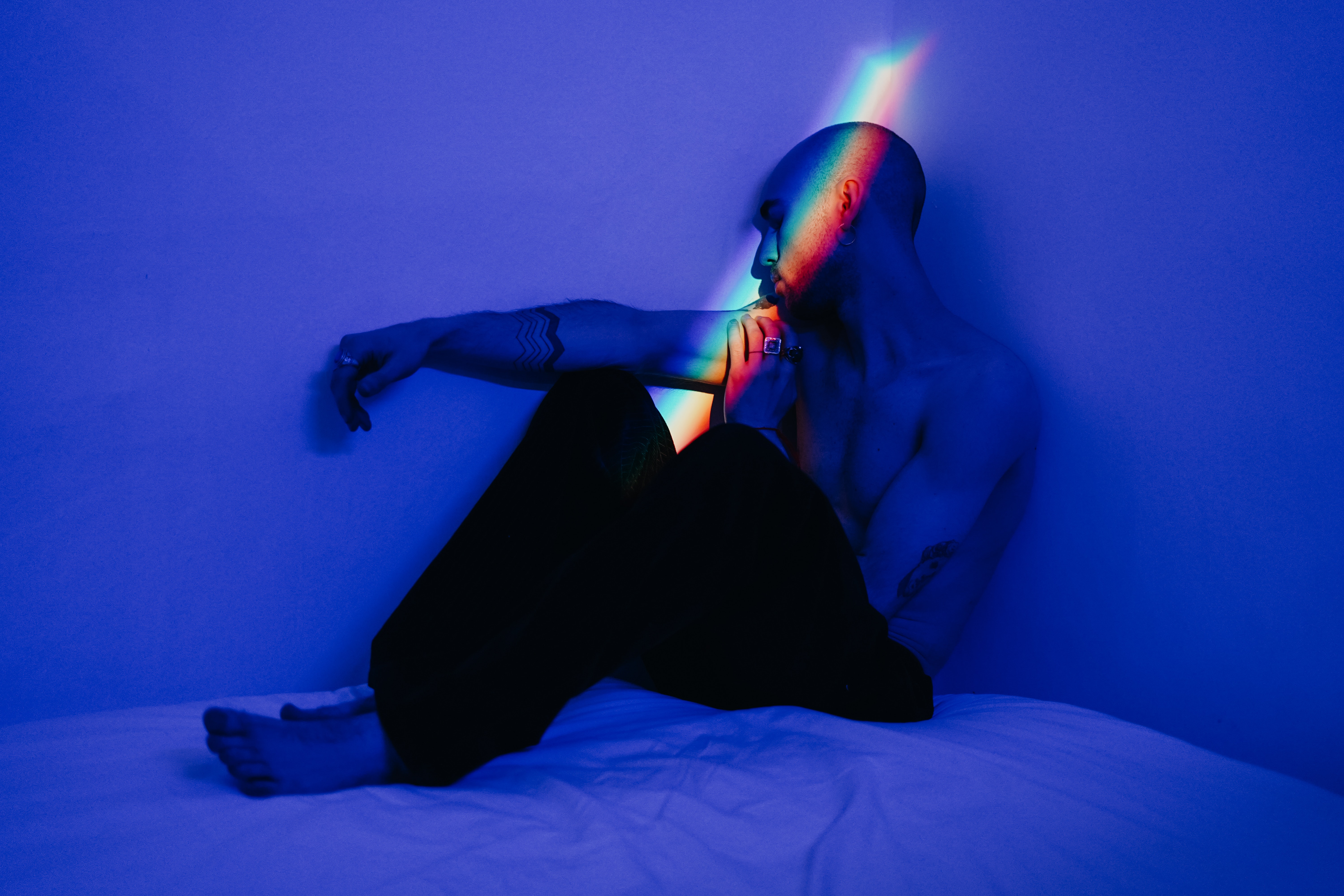 Masculine-presenting person sitting on a bed in the corner of a room with their head leaning against the wall and their right arm resting on their right knee. It looks dark - there is a blue hue across the image with one strip of light going across the person's face and arm. The strip of light is rainbow-coloured.