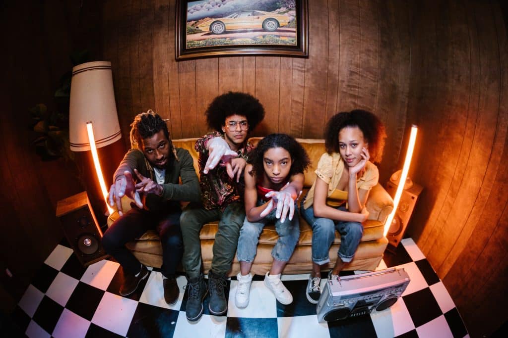 A fish-eye lens photo of 4 twenty-something year olds in a room sitting on a light brown couch looking and pointing at the camera. It's a diverse group and there are vintage speakers next to the couch on either side as well as a framed image of a yellow sports car hanging behind them.