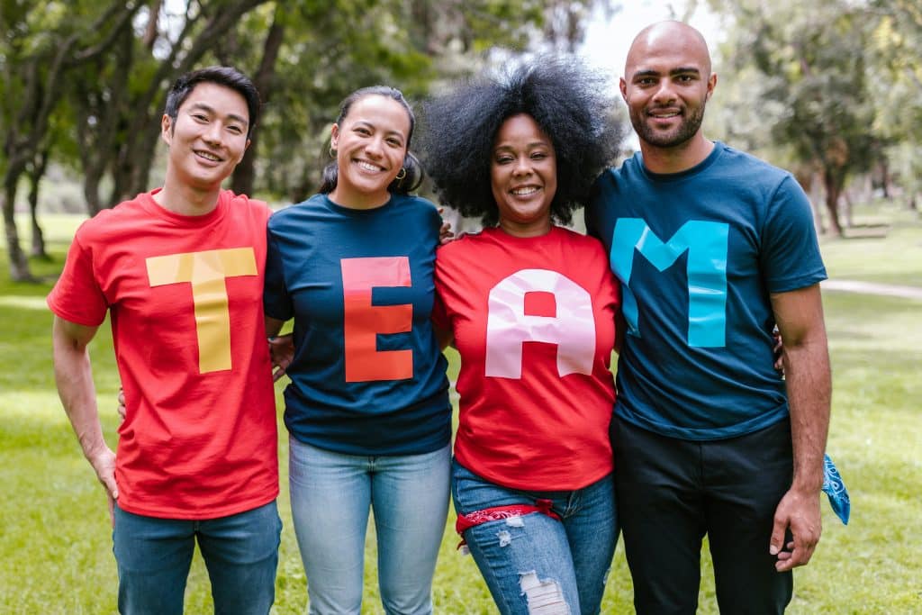 A photo of 4 diverse individuals standing next to each other side by side and arms around each other smiling and looking at the camera. They are wearing different coloured shirts but each person has a letter on the shirt and the 4 letters spell "TEAM" in capital letters. The background is blurred but obviously a park with lots of green grass and trees.