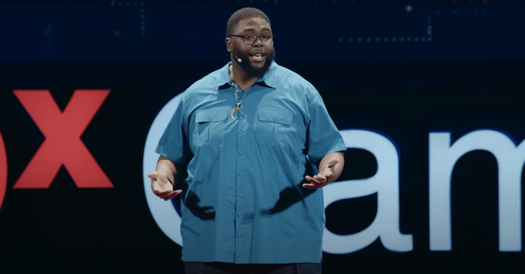 Anthony Abraham Jack, a middle-aged Black man, is wearing a short sleeve blue button up and gold necklace, is in the middle of delivering his TEDTalk on stage. The backdrop is zoomed in on the TEDxCambridge logo and showcases the "x" of the TEDx and the "Cam" of "Cambridge".