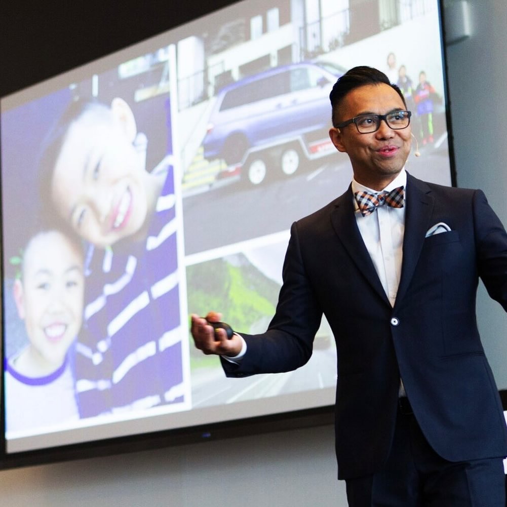 Alden Habacon, a middle-aged Filipino man dressed in a navy blue suit with a patterned bowtie, is on stage speaking to a crowd. He is smiling as a projector screen behind him showcases an image of 2 young boys (his sons) and other family photos.