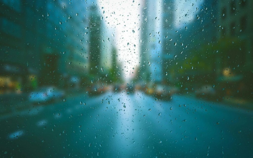 Raindrops in sharp focus on a window with a blurry city street and tall buildings in the background, evoking clarity emerging from noise.