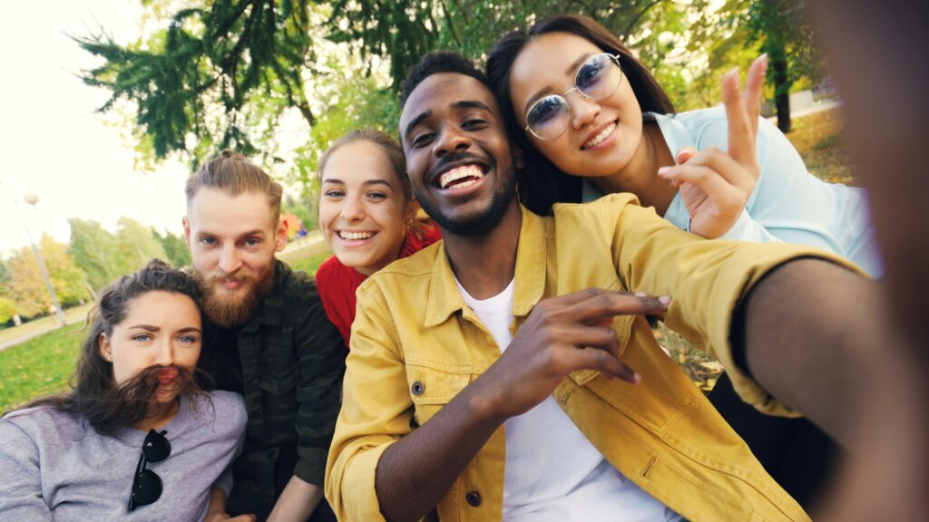 A diverse group of five smiling young adults taking a playful outdoor selfie together in a sunny park.
